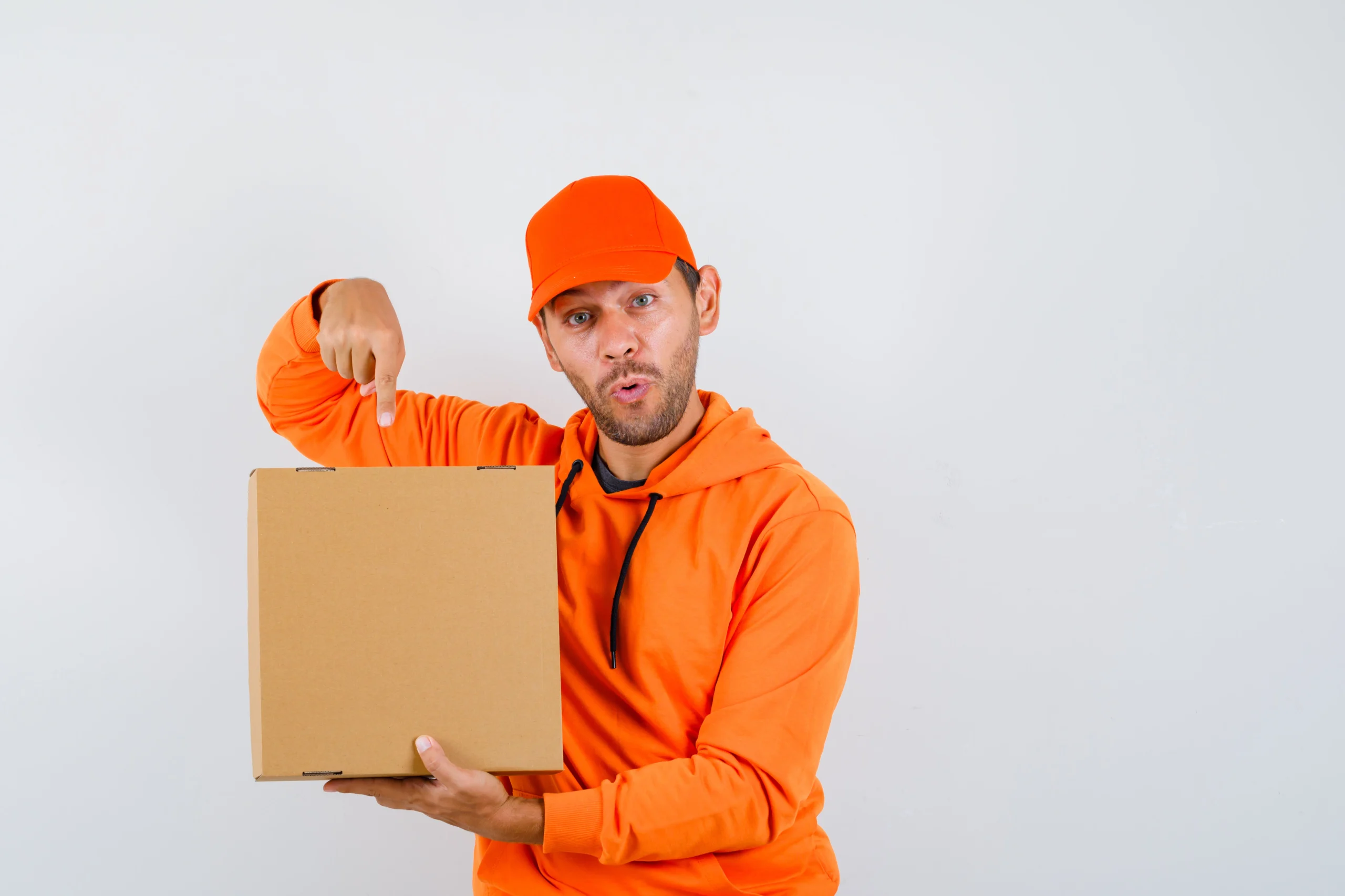 young delivery man pointing at cardboard box in hoodie, cap and looking wondered , front view.